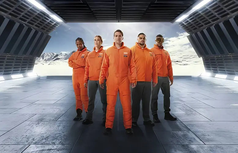 A group of five individuals dressed in orange spaceflight suits standing in a row inside a futuristic-looking structure with large windows showing a mountainous landscape, possibly on another planet. The spaceflight suits have badges, suggesting the individuals may be part of a space agency or team. To the right, there is a person wearing a pair of golden visor glasses. The room is illuminated by natural light, giving the scene an exploratory and adventurous atmosphere.